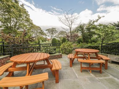 An outdoor patio with two wooden round picnic tables and benches surrounded by plants and trees at The Manor House in Scarborough
