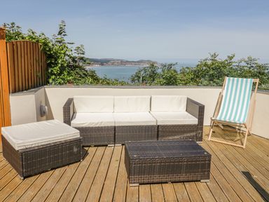 An outdoor patio with wicker seating including a sofa, ottoman, coffee table, and a striped deck chair with a sea view at Sea-Prize View in Old Colwyn