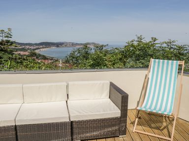 An outdoor seating area with a woven sofa and a striped deck chair overlooking a coastline at Sea-Prize View in Old Colwyn