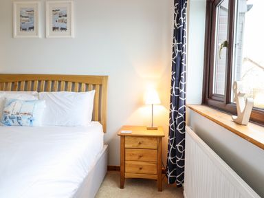 A bedroom with a wooden bed frame white bedding a wooden nightstand with a lamp and a window with curtains at Sea-Prize View in Old Colwyn