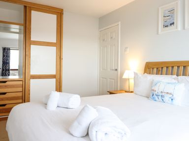 A bedroom with a bed with white linens wooden headboard rolled towels on bed wooden wardrobe and bedside table with lamp at Sea-Prize View in Old Colwyn