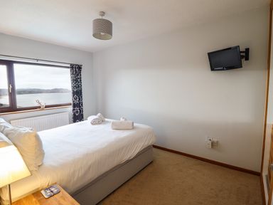 A bedroom with a bed with white linens a wooden nightstand a small wall-mounted tv and a window with a view of water and hills at Sea-Prize View in Old Colwyn
