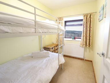 A small bedroom with a white metal bunk bed a window with curtains a small wooden drawer unit and a radiator at Sea-Prize View in Old Colwyn