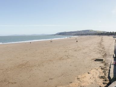 A sandy beach with a few people walking along the shore and a distant view of hills and houses at Sea-Prize View in Old Colwyn