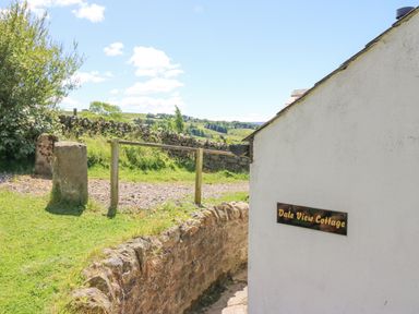 Exterior side of a white building with a Dale View Cottage sign and a stone wall with green fields in the background at Dale View Cottage in Allendale near Hexham