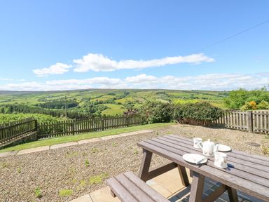 An outdoor wooden picnic table with teapot and cups on a gravel patio overlooking green hills and fields at Dale View Cottage in Allendale near Hexham