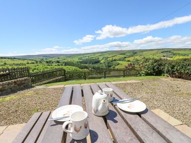 An outdoor wooden picnic table with a teapot mugs and plates overlooking a green fenced countryside at Dale View Cottage in Allendale near Hexham