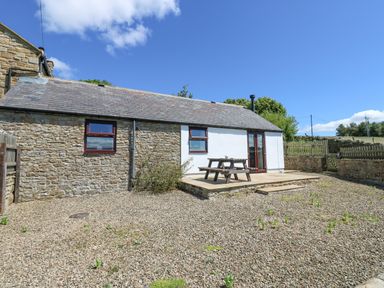 An outdoor area with a gravel ground, a wooden picnic table on a raised stone patio, and a small building with stone and white walls at Dale View Cottage in Allendale near Hexham