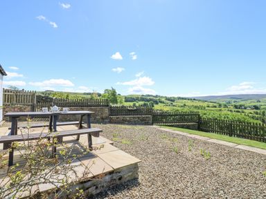An outdoor patio area with a wooden picnic table set with cups and teapot overlooking a fenced garden and green countryside at Dale View Cottage in Allendale near Hexham