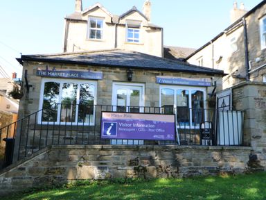 A stone building with signs for The Market Place Post Office gifts and Visitor Information with a railing in front