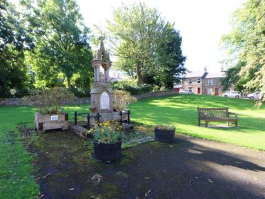 A small park with a stone monument, flower pots, a wooden bench, trees, and houses in the background at Dale View Cottage in Allendale near Hexham