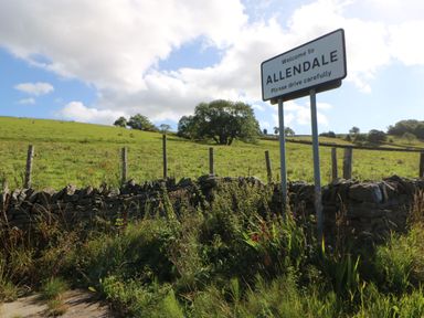 A countryside scene with a welcome to Allendale sign near a stone wall and green fields at Dale View Cottage in Allendale near Hexham