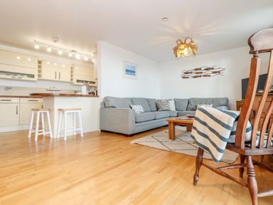 An open plan living room and kitchen with a grey sectional sofa, wooden coffee table, kitchen island with stools, and wooden chair with a striped blanket at Ocean Blue in Praa Sands
