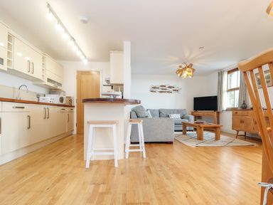 An open-plan kitchen and living room with wooden flooring white kitchen cabinets a grey sofa wooden stools a coffee table and a television at Ocean Blue in Praa Sands