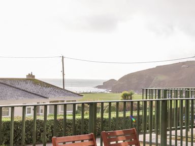 A balcony with wooden chairs overlooking a lawn a house sea and hills at Ocean Blue in Praa Sands
