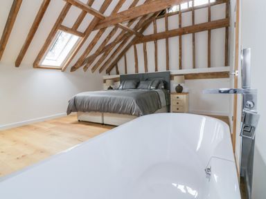 A bedroom with a bed and bedside tables under a slanted wooden beam ceiling and a bathtub in the foreground at Grange Barn in Gedding near Felsham