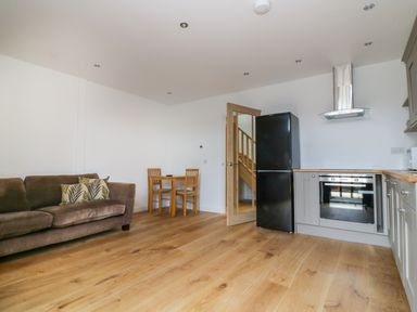 A room with a brown sofa, wooden dining table with two chairs, black refrigerator, built-in oven, and wooden floor at Grange Barn in Gedding near Felsham