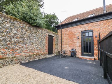 An outdoor courtyard with a gravel and asphalt ground a stone wall with a wooden door a brick building with a glass door black fence two chairs a table and a barbecue grill at Grange Barn in Gedding near Felsham