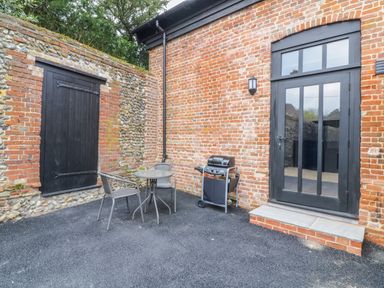 An outdoor patio area with a small round table and two chairs a barbecue grill and a black door in a brick wall at Grange Barn in Gedding near Felsham