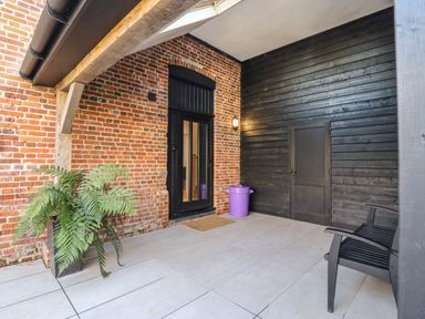 A covered outdoor patio area with brick and black wooden walls a black door a black chair a potted plant and a purple trash bin at Grange Barn in Gedding near Felsham