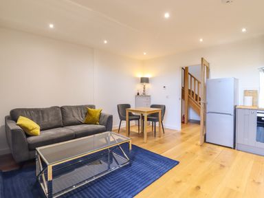 A living room with a grey sofa with yellow cushions a glass coffee table a blue rug a small dining table with two chairs a lamp on a cabinet a refrigerator and wooden stairs at Grange Barn in Gedding near Felsham