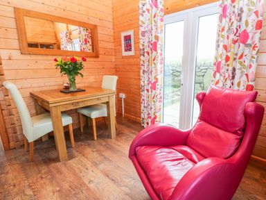 A dining area with a wooden table white chairs a red leather armchair and floral curtains by glass doors at Borah Lodge in Mousehole