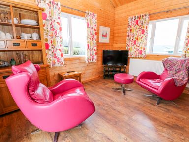A wooden living room with two red chairs a matching footstool a wooden cabinet floral curtains and a television at Borah Lodge in Mousehole