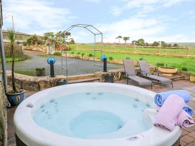 An outdoor hot tub with towels beside two lounge chairs and a garden with a fence and trees at Meadowside in Mousehole
