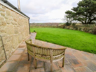 A wooden bench on a stone patio next to a stone building with a green lawn and trees in the background at Meadowside in Mousehole