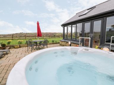 An outdoor patio with a hot tub a table and chairs with a red umbrella and a grassy field behind at Meadowside in Mousehole