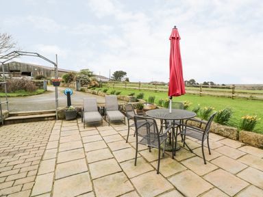 An outdoor patio with a table and four chairs with a closed red umbrella and two lounge chairs on stone tiles at Meadowside in Mousehole