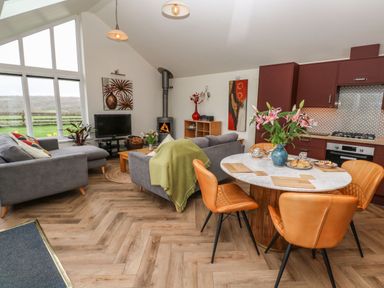 An open plan living area with grey sofas a wood burning stove a round dining table with orange chairs and a kitchen with red cabinets at Meadowside in Mousehole
