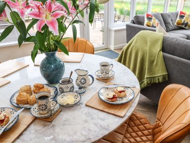A dining table set with cups of coffee scones and cream in a room with a sofa and large windows at Meadowside Mousehole