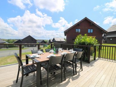 An outdoor dining table with chairs on a wooden deck overlooking houses and greenery at Willow Retallack in Retallack near St Columb Major