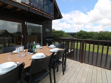 An outdoor dining table set with bowls, glasses, and bottles on a wooden deck with a railing at Willow, Retallack in Retallack near St Columb Major