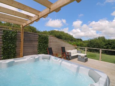 An outdoor deck with a hot tub wooden pergola chairs a sofa and a table with greenery and a blue sky at Willow Retallack in Retallack near St Columb Major