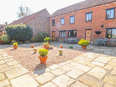 A paved courtyard with potted plants and outdoor metal tables and chairs in front of a brick building at The Dairy Barn in Toftrees near Fakenham