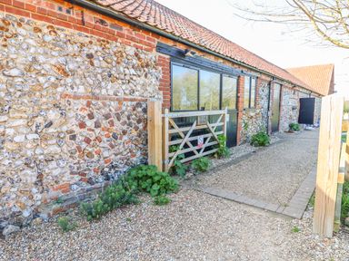An exterior view of a stone and brick barn with a gravel pathway and a wooden gate at The Dairy Barn in Toftrees near Fakenham
