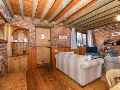 A living room with wooden beams on the ceiling a wooden door a window with curtains striped sofas a bookshelf and a TV at The Dairy Barn in Toftrees near Fakenham