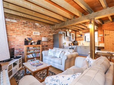 An open living room and kitchen with exposed wooden beams brick walls and light grey sofas at The Dairy Barn in Toftrees near Fakenham