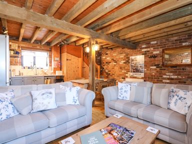 A living room with two sofas and cushions a wooden coffee table with magazines a dining table with chairs a kitchen area with cabinets and a window exposed wooden beams and a brick wall with a poster at The Dairy Barn in Toftrees near Fakenham