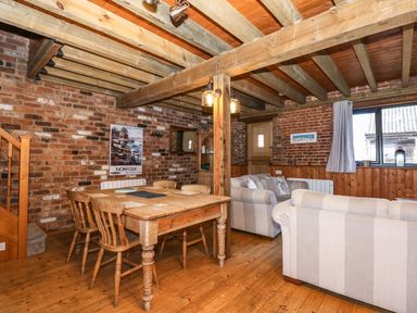 A dining table with chairs and two sofas in a room with exposed brick walls and wooden beams at The Dairy Barn in Toftrees near Fakenham