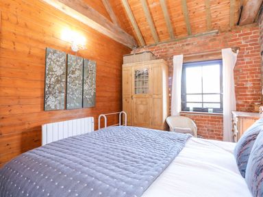A bedroom with wooden and brick walls a bed with a quilt a wardrobe a wicker chair and a window with curtains at The Dairy Barn in Toftrees near Fakenham