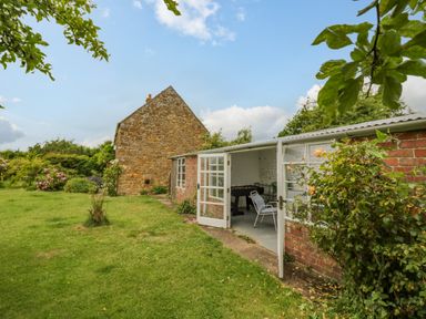 A garden with a lawn and flowers next to a brick outbuilding with open doors showing a chair and foosball table Priors Mead Claydon near Cropredy