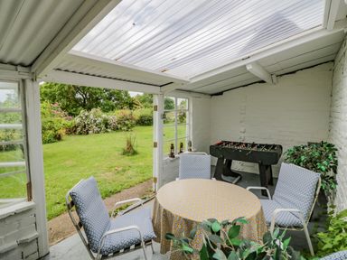 A conservatory with a round table covered with a patterned tablecloth surrounded by four cushioned chairs and a foosball table at Priors Mead in Claydon near Cropredy