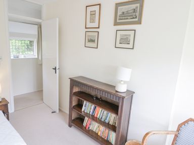 A room with a wooden bookshelf filled with books a white lamp on top four framed pictures on the wall an open door and a chair at Priors Mead in Claydon near Cropredy