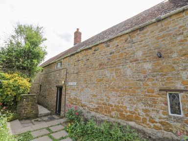 An exterior view of a stone building with a chimney a small rectangular window and plants along a stone path at Priors Mead Claydon near Cropredy