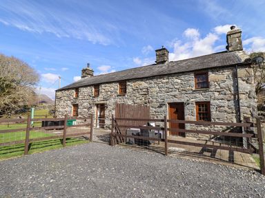 A stone cottage with three chimneys and wooden doors behind a fenced yard at Cwm Yr Afon Mawr in Llanbedr near Harlech