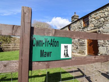 A wooden gate with a green sign showing a dog and the text Cwm Yr Afon Mawr in front of a stone building with wooden door and windows at Cwm Yr Afon Mawr Llanbedr near Harlech