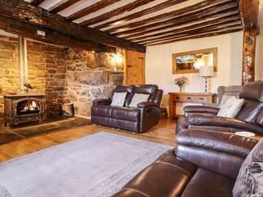 A living room with leather sofas a stone fireplace wooden ceiling beams and a wooden side table with a lamp and flowers at Cwm Yr Afon Mawr in Llanbedr near Harlech